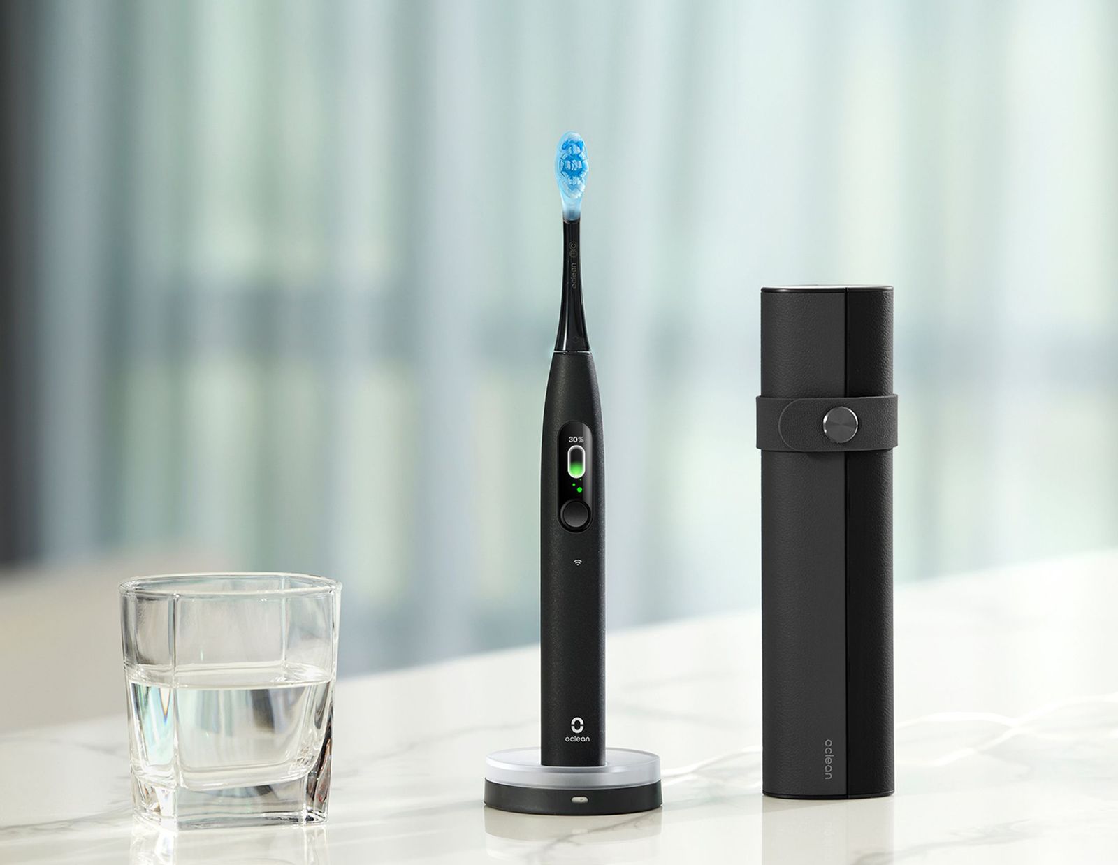 Black electric toothbrush sitting on a marble counter beside a glass of water and a black carrying case