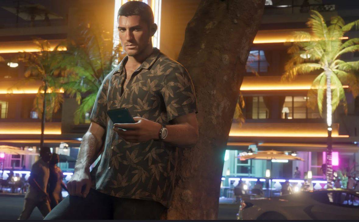 One of two protagonists in GTA 6, Jason, holds a phone as he leans against a tree. He's wearing a patterned shirt with leaves as the city nightlife bustles in the background. 